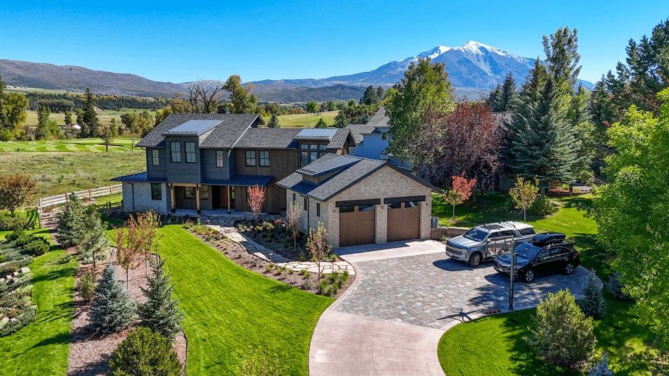 Aerial view of a two-story mountain home with a stone garage, landscaped lawn and a snow-capped peak behind