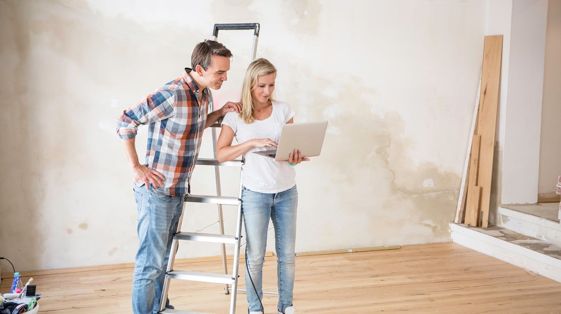 Couple standing by a stepladder looking at a laptop while planning a home renovation