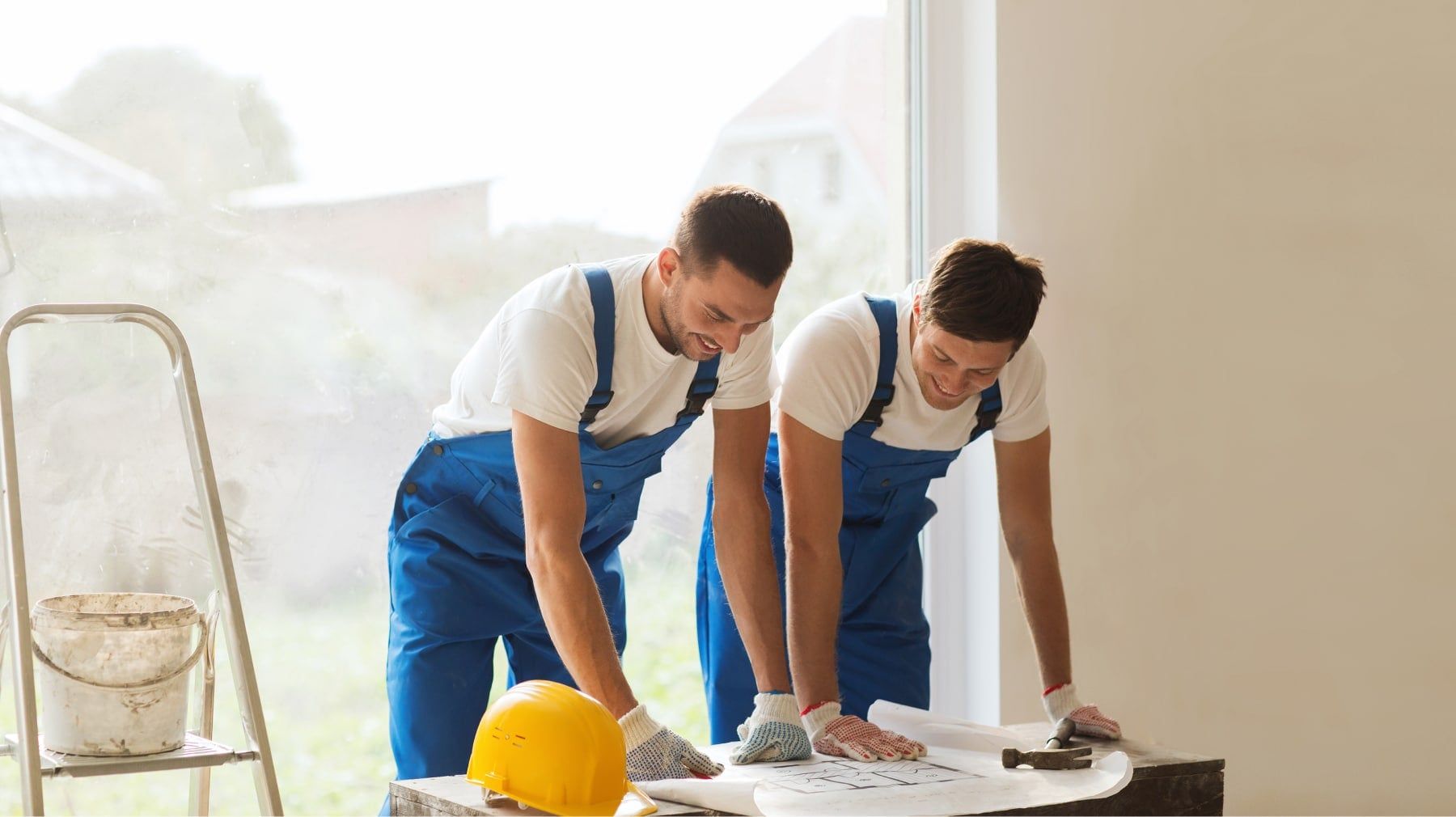 Two remodelers in overalls reviewing plans next to a stepladder and hard hat