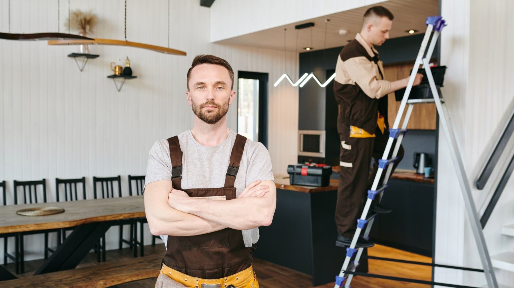 Contractor with arms crossed in a kitchen while a coworker works on a ladder