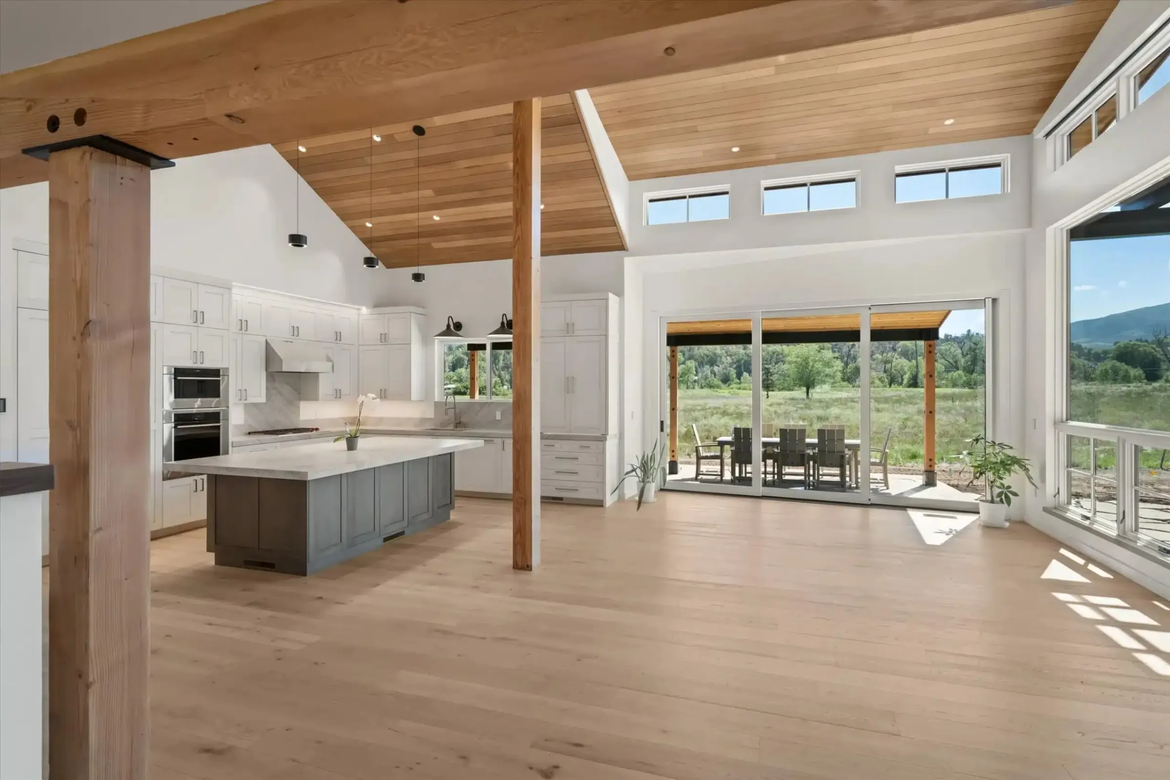 Vaulted great room with a wood ceiling, timber posts and an open white kitchen facing a covered patio