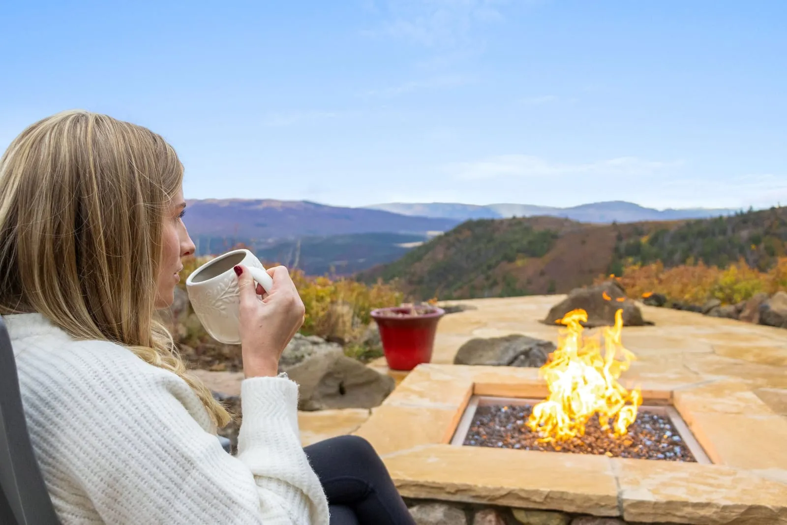 Woman holding a mug beside a stone fire pit on the new patio, with autumn hills beyond