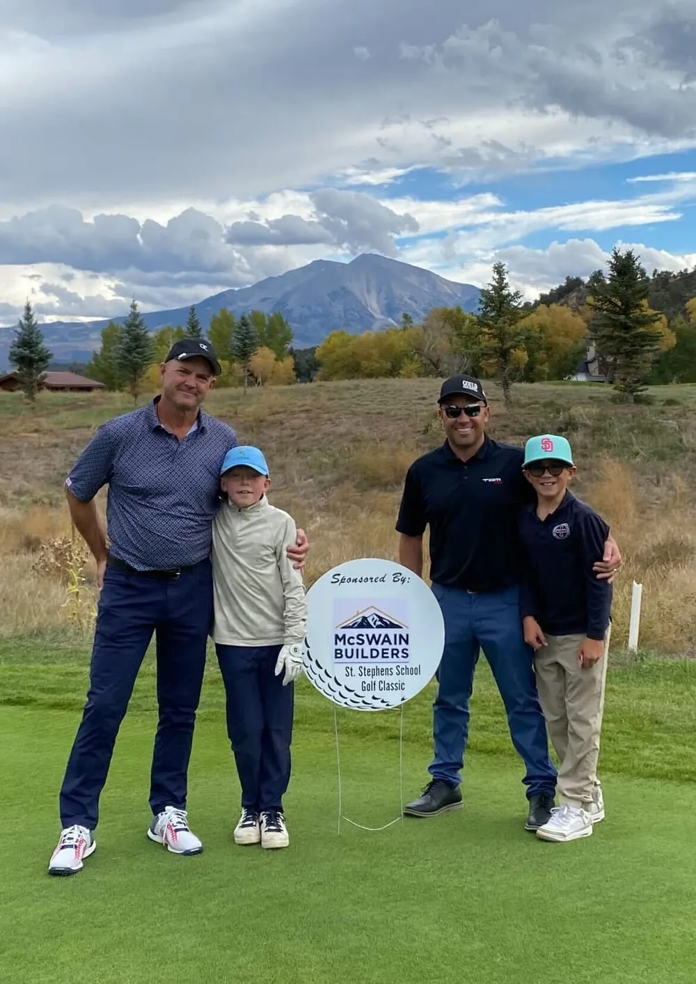 Two golfers and two boys on the green beside a sign reading Sponsored by McSwain Builders, St. Stephens School Golf Classic