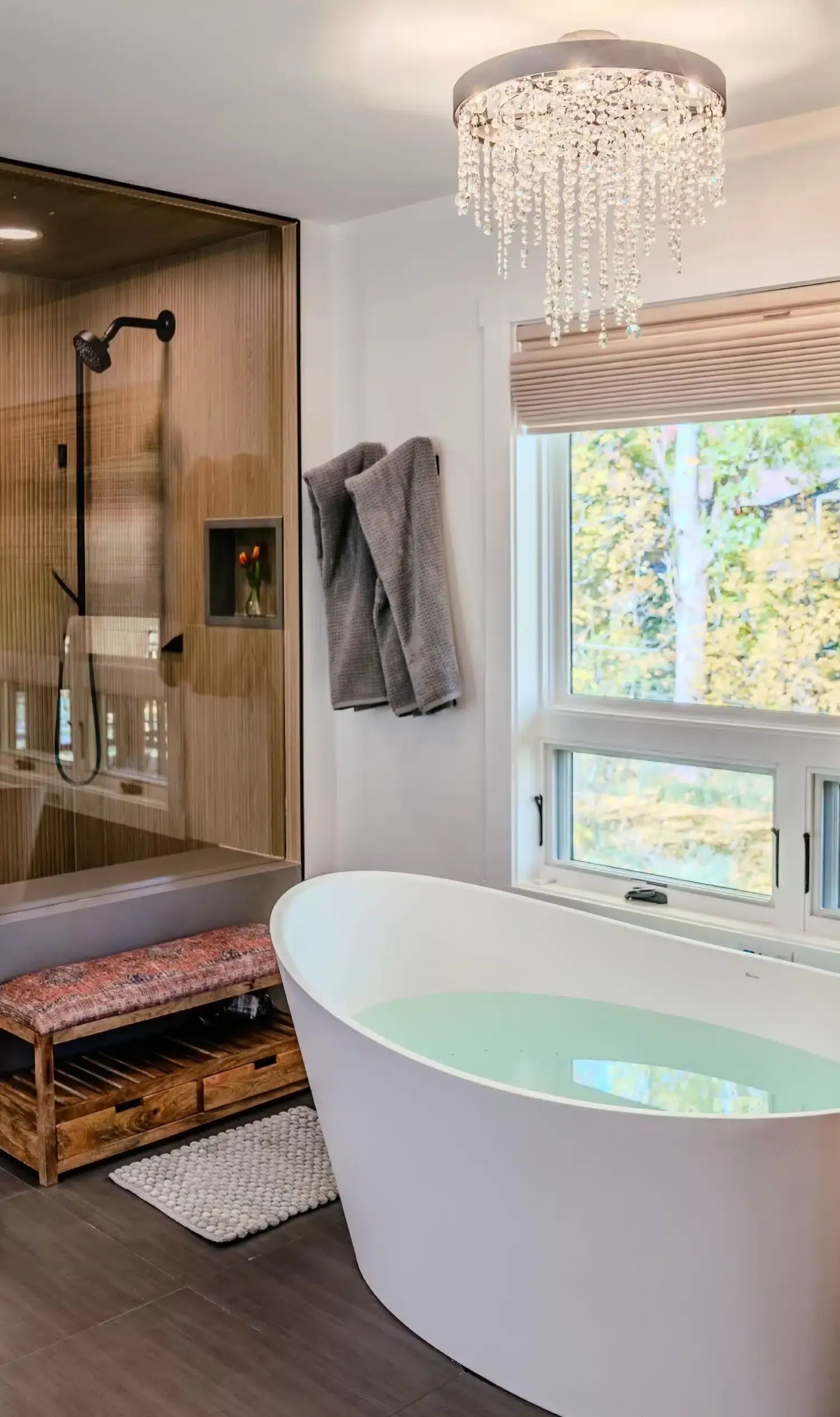 Freestanding soaking tub under a crystal chandelier beside a wood-paneled shower