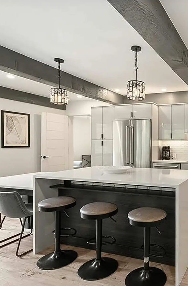 White waterfall island with black bar stools under gray ceiling beams and crystal pendants